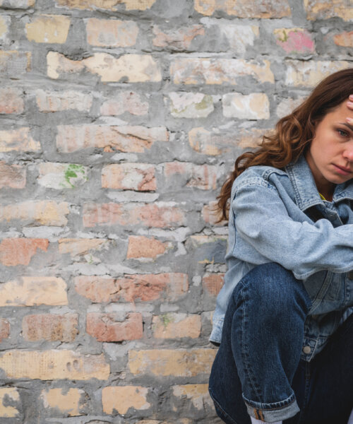 Alone stressed and tired urban woman sits next to the brick wall on the street in city.